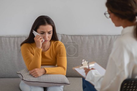 Photo for A young woman in a somber mood consults with a therapist who is taking notes, depicting a therapy session - Royalty Free Image