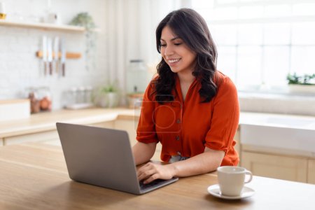 Foto de Una mujer de Oriente Medio se sienta cómodamente en casa, interactuando con su portátil. Ella tiene una expresión feliz, representando a un zoómero que utiliza la tecnología - Imagen libre de derechos