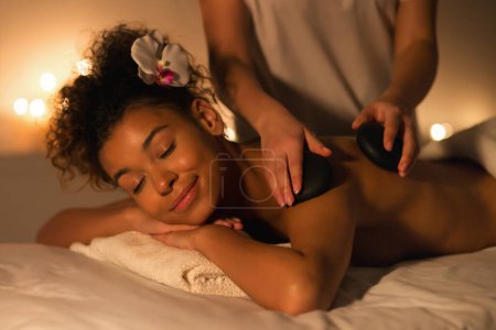 Photo for Serene image of hot stone massage therapy for african american woman, highlighting well-being and relaxation techniques in a peaceful spa environment - Royalty Free Image