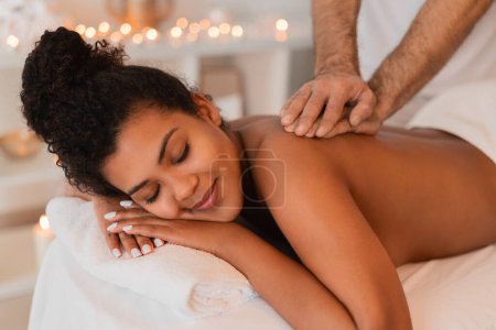 Téléchargez les photos : Femme afro-américaine souriant de contentement alors qu'elle reçoit un massage du dos dans un spa avec une ambiance chaleureuse - en image libre de droit