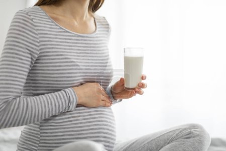 Téléchargez les photos : Une dame européenne à la maison berce sa bosse de bébé tout en tenant un verre nutritif de lait, soulignant l'importance de la santé pendant la grossesse - en image libre de droit