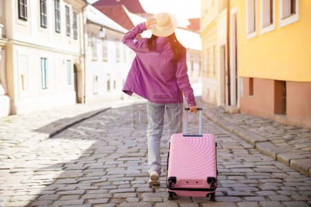 Téléchargez les photos : Photo en plein air d'une étudiante millénaire marchant avec une valise rose dans le paysage urbain européen, dépeignant des idéaux de liberté et de nouvelles expériences - en image libre de droit