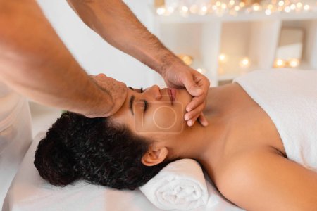 A skilled masseur provides a relaxing face massage to a client African American lady in a tranquil spa setting with dim lighting and candles