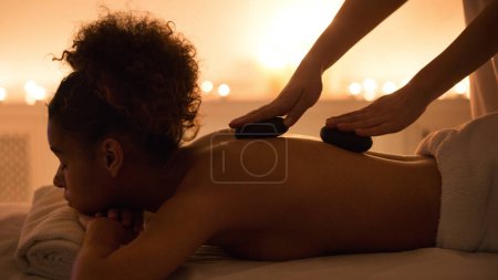 Téléchargez les photos : Une image sereine capturant un moment de détente, la femme afro-américaine profite d'un massage aux pierres chaudes dans un cadre spa faiblement éclairé La chaleur et les soins sont palpables - en image libre de droit