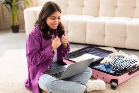 Téléchargez les photos : Cette image montre une femme du Moyen-Orient confirmant avec enthousiasme les plans de voyage sur son ordinateur portable à la maison, avec une valise à côté d'elle - en image libre de droit