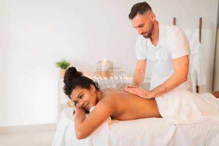 Photo for A professional masseur provides a back massage to a relaxed African American woman lying on a massage table in a spa - Royalty Free Image