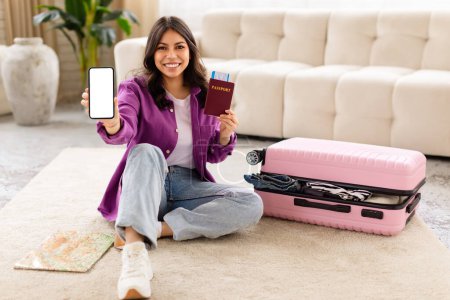 Téléchargez les photos : Une joyeuse femme du Moyen-Orient s'assoit sur le sol, tenant un smartphone et un passeport, avec une valise ouverte et une carte à proximité - en image libre de droit
