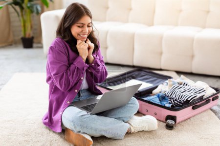 A muslim woman appears joyful while looking at her laptop at home, with a suitcase suggesting travel readiness