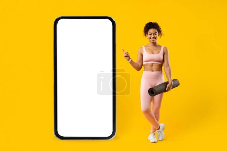 Foto de Una joven afroamericana sonriente en ropa deportiva sostiene una esterilla de yoga y señala una pantalla de teléfono inteligente en blanco sobre un fondo amarillo brillante - Imagen libre de derechos