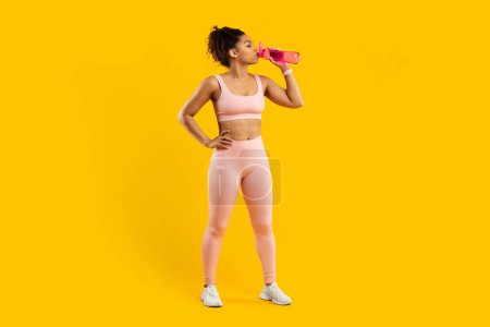 A fit african american woman in pink sportswear pauses to hydrate with a water bottle against a vibrant yellow background, showcasing health and active lifestyle