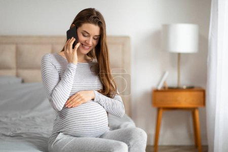 Foto de Una mujer embarazada sonriente se ve comunicándose en un teléfono inteligente mientras toca suavemente su vientre, sentado cómodamente en un acogedor dormitorio - Imagen libre de derechos