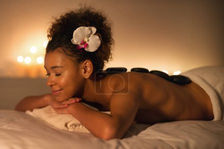 Photo for A young african american woman lies face down on a spa bed with hot stones on her back, indicating a luxuriating spa treatment in a serene ambiance - Royalty Free Image