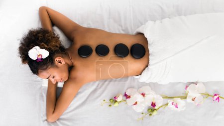 Photo for A peaceful african american lady enjoys a spa massage featuring black stones and orchids, embodying tranquility and harmony in a serene spa setting, top view - Royalty Free Image