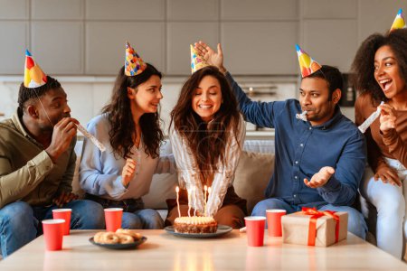 Foto de Jóvenes amigos multirraciales alegres celebran un cumpleaños con torta y sombreros festivos en casa, capturando un momento de felicidad - Imagen libre de derechos