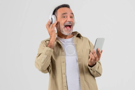 Téléchargez les photos : Vieil homme isolé sur fond blanc, interagit joyeusement avec la musique à l'aide d'écouteurs blancs et d'un smartphone, montrant une adaptabilité technologique des aînés - en image libre de droit