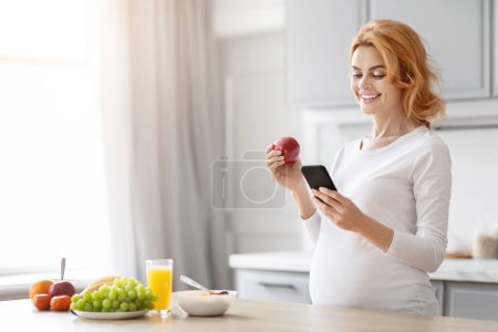 Téléchargez les photos : Femme enceinte européenne vérifie un téléphone portable, avec un accent sur la nutrition, debout dans une cuisine avec des fruits à proximité - en image libre de droit