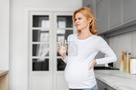 Foto de Una mujer embarazada con una camisa blanca está de pie en una cocina, sosteniendo un vaso de agua y tocándose la espalda con una expresión dolorosa - Imagen libre de derechos