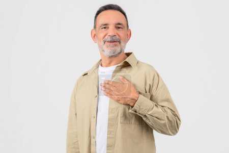 A senior man portrays sincerity and warmth with a hand-on-heart gesture, wearing casual attire isolated on a white background