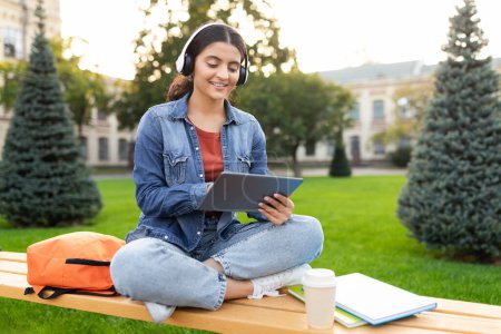 This serene image shows an Indian lady using a tablet while sitting on a bench, an embodiment of modern education and technology on a lush green campus, showcasing the life of an engaged zoomer
