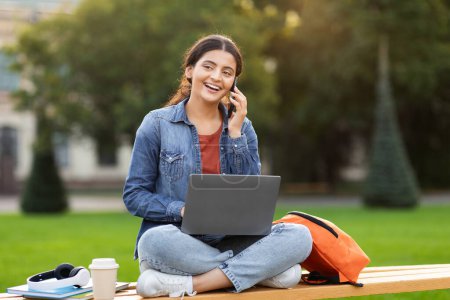 Photo for Casual indian lady speaks on phone at a campus, conveying connectivity and modern education with her relaxed posture and laptop - Royalty Free Image