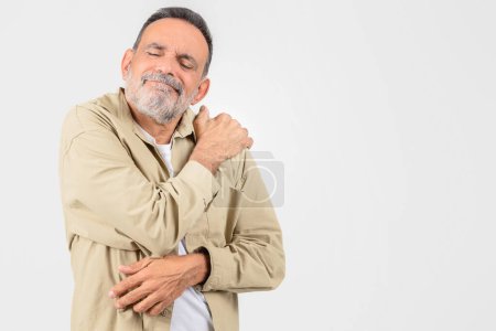 Téléchargez les photos : Un homme âgé soulagé touche son épaule avec un sourire content, isolé sur blanc, exprimant éventuellement la guérison de la douleur ou de l'inconfort, copier l'espace - en image libre de droit