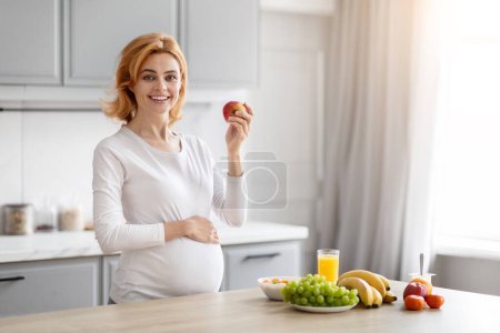 Photo for A joyful pregnant European lady stands in the kitchen holding an apple, symbolizing healthy nutrition choices during pregnancy - Royalty Free Image