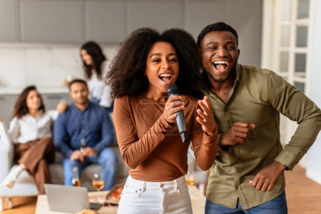 Téléchargez les photos : Des amis multiraciaux partagent un microphone, chantant joyeusement avec des rires et des expressions amusantes dans un cadre confortable à la maison - en image libre de droit