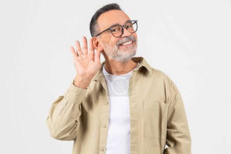 Ein Großvater im Ruhestand, isoliert auf Weiß, ringt ums Hören und gestikuliert mit der Hand hinter dem Ohr, was auf Hörschwierigkeiten hindeutet, die bei alten Menschen üblich sind.