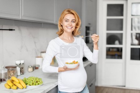 Foto de Alegre señora embarazada europea en un entorno de cocina sonríe mientras sostiene un recipiente nutritivo, haciendo hincapié en la nutrición de la cocina - Imagen libre de derechos