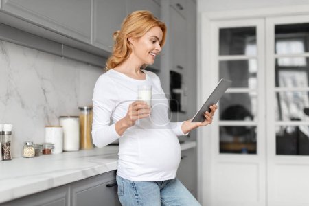 Photo for European pregnant lady casually enjoying milk while browsing on a tablet in her kitchen, set against a theme of digital age and prenatal nutrition - Royalty Free Image