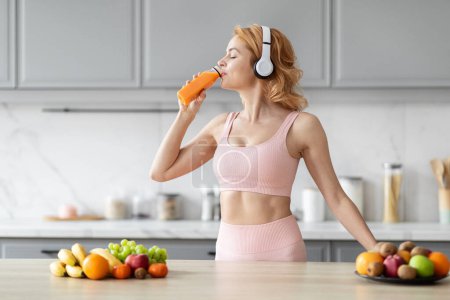 Photo for In a modern kitchen, a sporty european lady snacking on a carrot represents her conscious nutrition choices after workout - Royalty Free Image