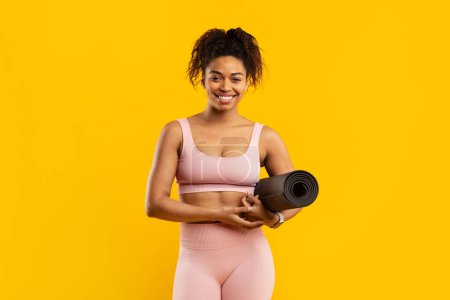 Téléchargez les photos : Une jeune femme afro-américaine heureuse et en forme en vêtements de sport tient un tapis de yoga, prêt pour une séance d'entraînement, sur un fond jaune vif - en image libre de droit