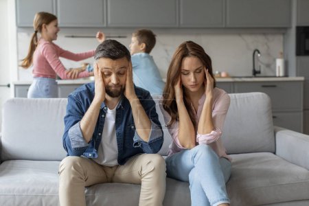 Photo for A stressed couple sits on a couch, looking exhausted, while their two children fight in the background of a modern home - Royalty Free Image