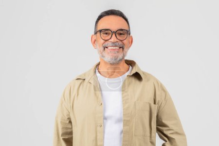 Ein fröhlicher älterer Mann mit Bart und Brille posiert in einem beigen Hemd vor grauem Hintergrund und strahlt Zuversicht und Glück aus.