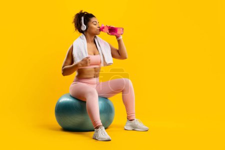Téléchargez les photos : Femme de fitness afro-américaine assoiffée prend une pause pour s'hydrater alors qu'elle est assise sur une balle de gym, isolée sur un fond jaune vif - en image libre de droit