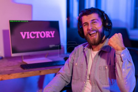 A passionate millennial guy fist pumps in front of a VICTORY screen at home, illustrating the addictive excitement of gaming