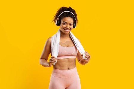 Photo for A joyful african american lady in fitness gear wearing headphones and a towel enjoys a workout break, isolated on yellow - Royalty Free Image