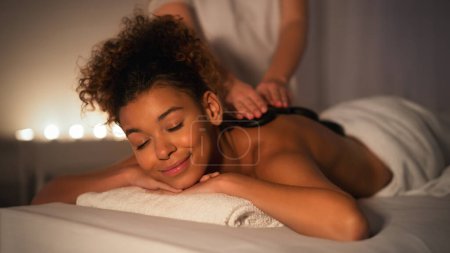 Foto de Capturada en un ambiente suavemente iluminado, una dama afroamericana se muestra tranquila durante una sesión de masaje de spa centrada en la relajación y el bienestar. - Imagen libre de derechos