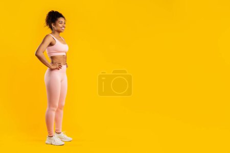 Eine durchtrainierte afrikanisch-amerikanische Frau posiert in Fitnessbekleidung, die Hände auf den Hüften, isoliert auf gelbem Hintergrund, Kopierraum