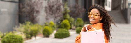 Foto de Retrato de la joven afroamericana sonriente en casual y gafas con bolsa con compras, mirando a la cámara en la calle de la ciudad, espacio de copia. Gran oferta y anuncio, shopaholic disfrutar de compras y venta - Imagen libre de derechos