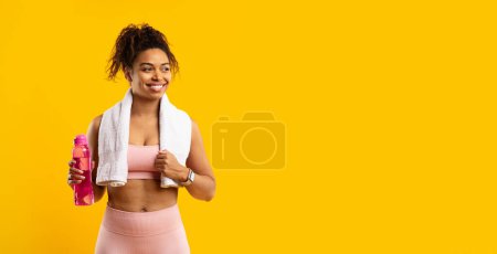 Téléchargez les photos : Femme afro-américaine radieuse en tenue de fitness tenant une bouteille d'eau rose, souriant avec une serviette autour du cou sur un fond jaune - en image libre de droit