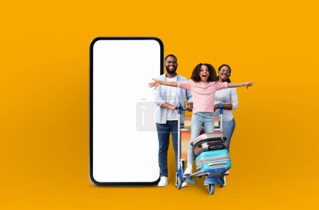 Téléchargez les photos : Une famille afro-américaine joyeuse avec des valises à côté d'un grand écran de téléphone, représentant des applications de vacances en famille et des offres en ligne d'une manière numérique isolée - en image libre de droit
