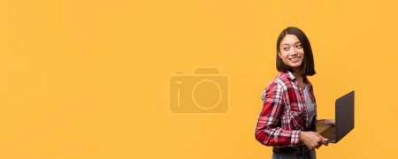 Foto de Una joven mujer asiática en una camisa a cuadros sostiene alegremente un portátil con un amplio espacio de copia sobre un fondo amarillo vivo - Imagen libre de derechos