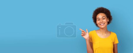 Téléchargez les photos : Femme afro-américaine souriante dans un haut jaune pointant vers l'espace vide sur un fond bleu, ce qui en fait une bannière web engageante ou une maquette - en image libre de droit