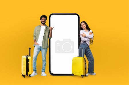 Téléchargez les photos : Couple indien confiant se tient à côté d'un smartphone géant avec un écran vide, approprié pour les affichages d'applications de voyage, avec une toile de fond jaune symbolisant la joie - en image libre de droit