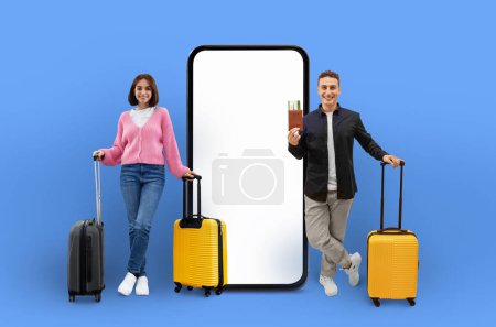 Photo for A young couple with luggage enthusiastically presents a large smartphone with a blank screen, suited for travel applications, set against a vibrant blue backdrop - Royalty Free Image