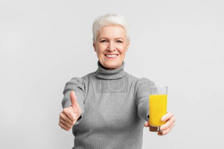 Cheerful elderly european woman gives a thumbs-up while holding orange juice emphasizing healthy s3niorlife