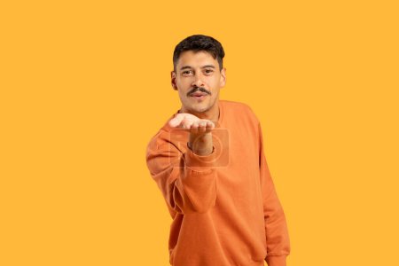 Ein Millennial-Mann mit Schnurrbart pustet amüsant einen Kuss in die Kamera, vor einer isolierten, leuchtend orangen Kulisse