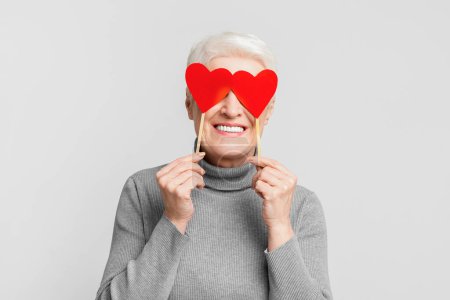 Une femme européenne âgée moderne couvrant ses deux yeux avec des bâtons rouges en forme de coeur et souriant sur fond gris