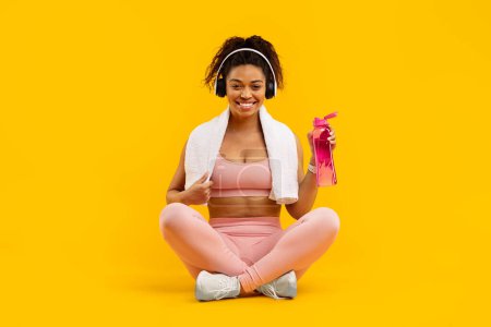 Eine afrikanisch-amerikanische Dame in Fitnesskleidung sitzt im Schneidersitz und lächelt mit einer Wasserflasche, isoliert auf gelb